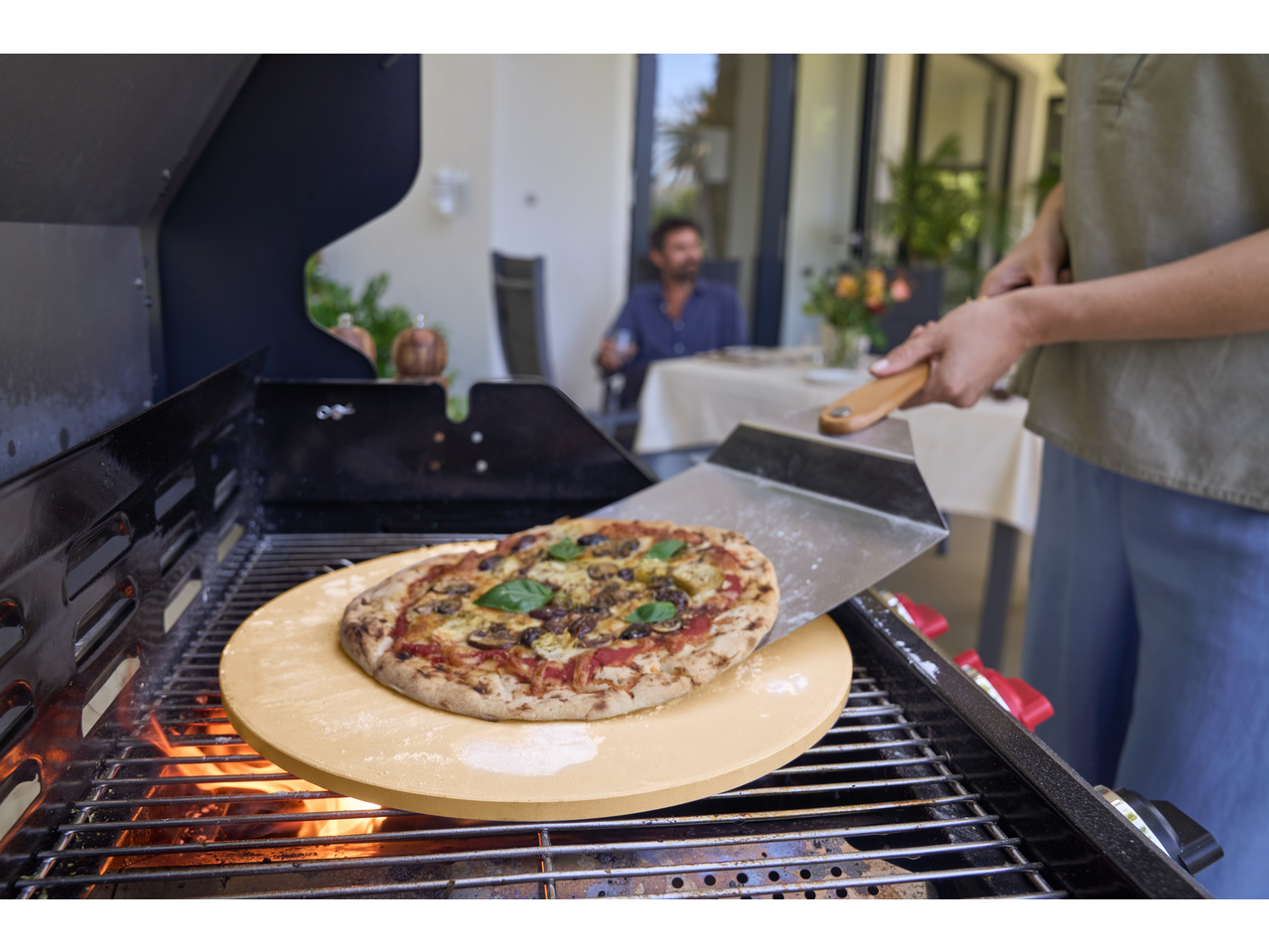 Grillmeister Pizza Stone with a pizza being placed on a grill, with a person in the background.