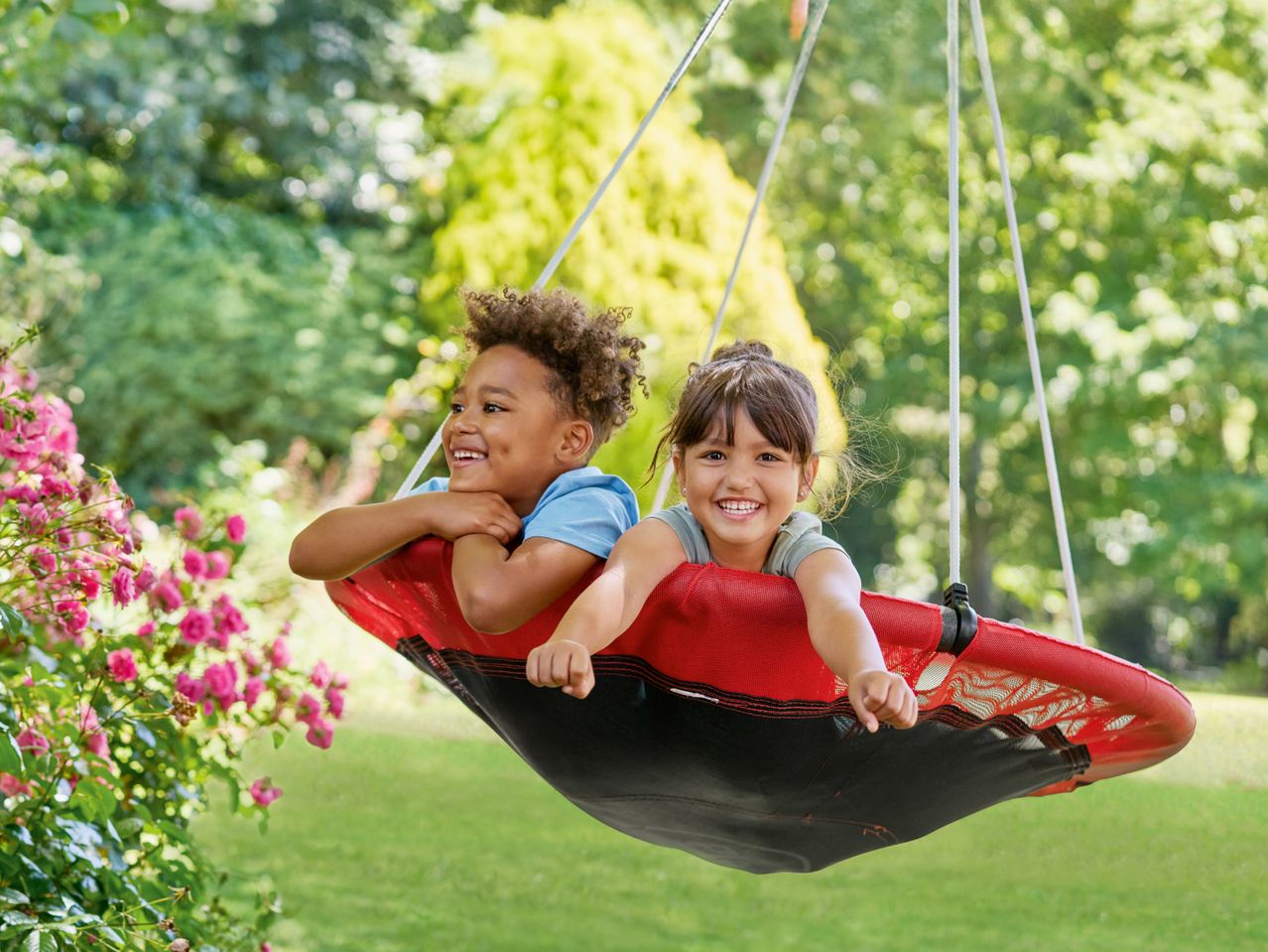 Zwei lächelnde Kinder in einer roten Nestschaukel im Garten.