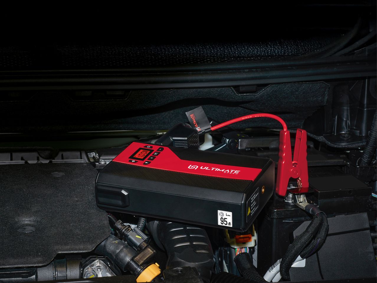 A red and black jump starter connected to a car battery in an engine bay.