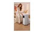 Woman sitting on a beige ottoman, adjusting a white portable air conditioner in a cozy room.