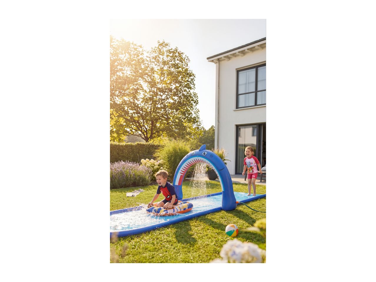 Two children playing on a shark-themed water slide in a sunny garden.