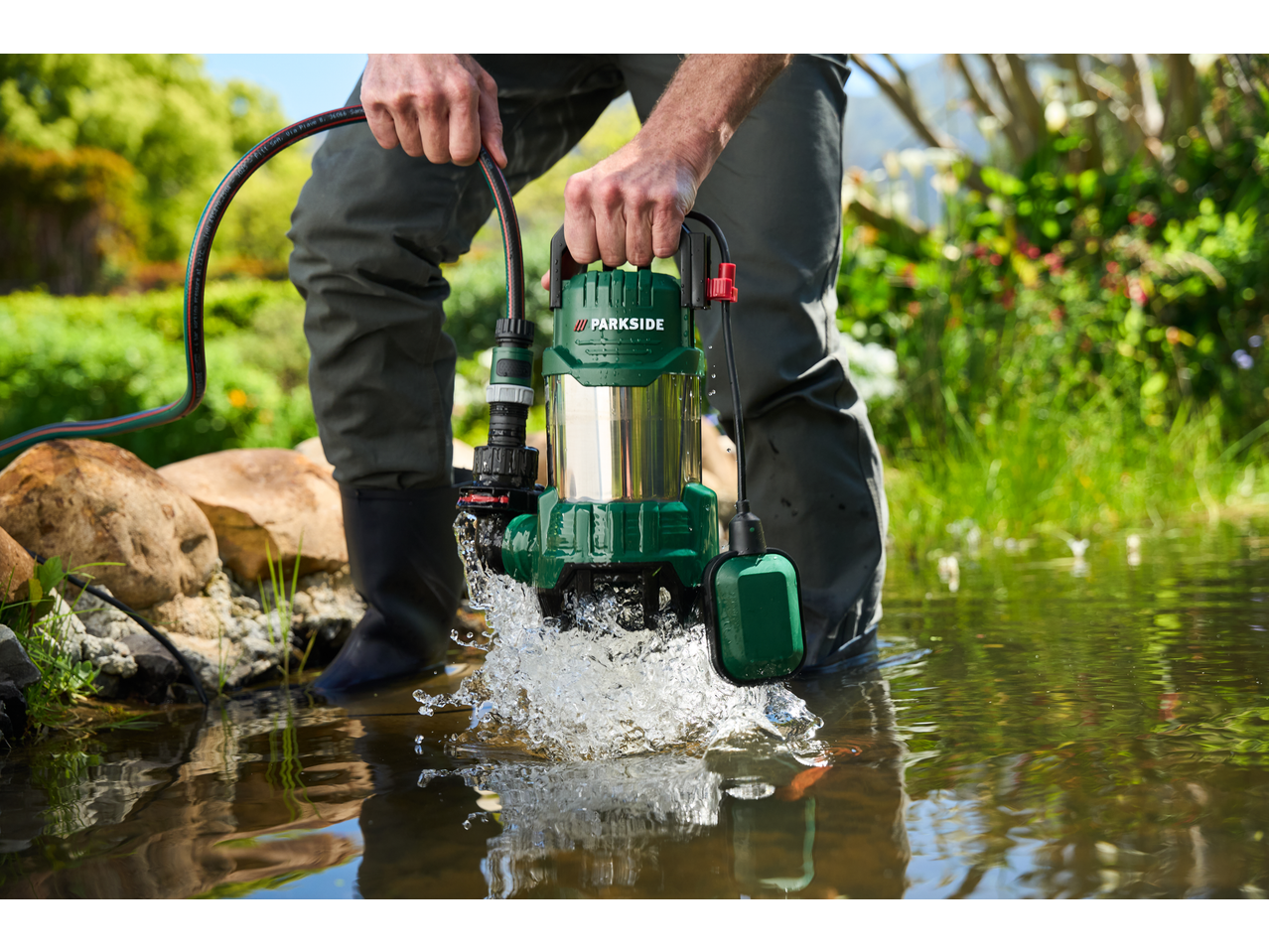 A person in waders places a green submersible pump into a pond, causing water to splash.