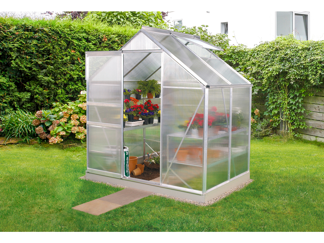 Vitavia Greenhouse in a garden, filled with potted plants and gardening tools.