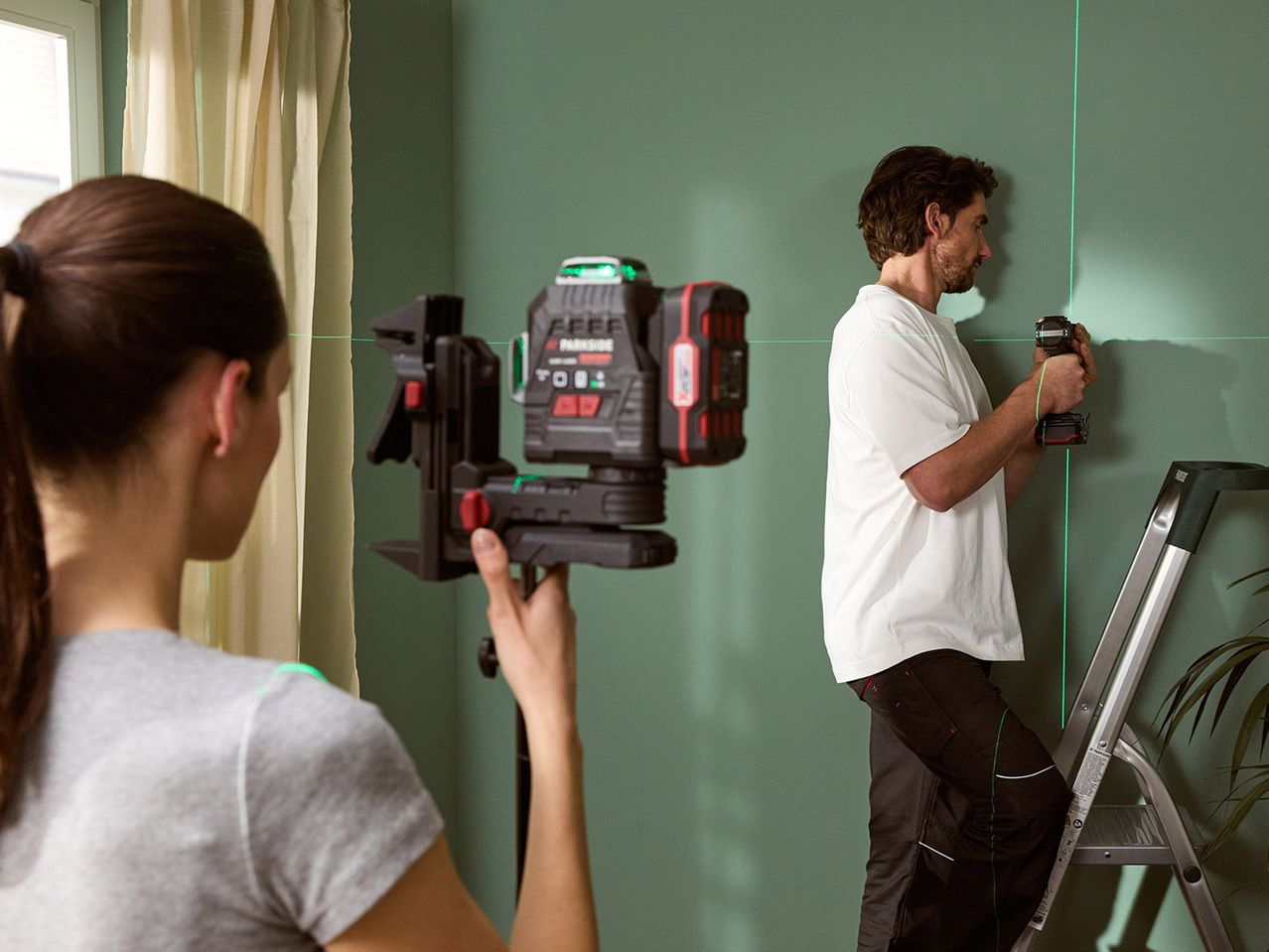 Μια γυναίκα χρησιμοποιεί ένα λέιζερ Bosch για να βοηθήσει έναν άνδρα να βάψει έναν τοίχο.