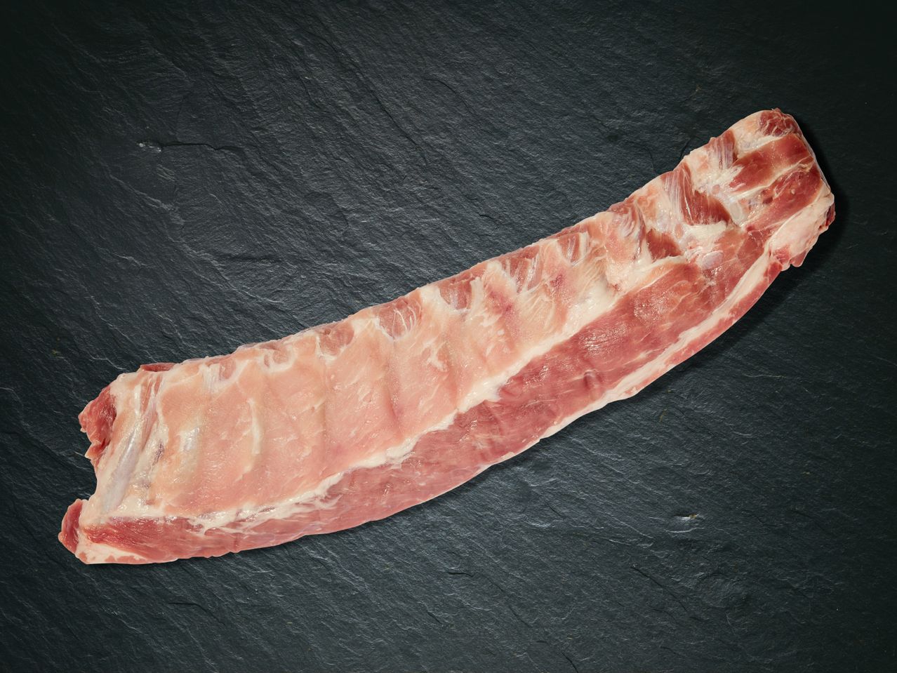 Ein Stück rohe Schweinerippchen auf einer schwarzen Schieferplatte.