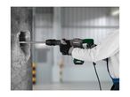 Person using a hammer drill to break concrete, with a blurred industrial background.