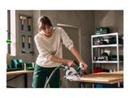 Woman using a circular saw to cut wood in a workshop, with tools on the wall.