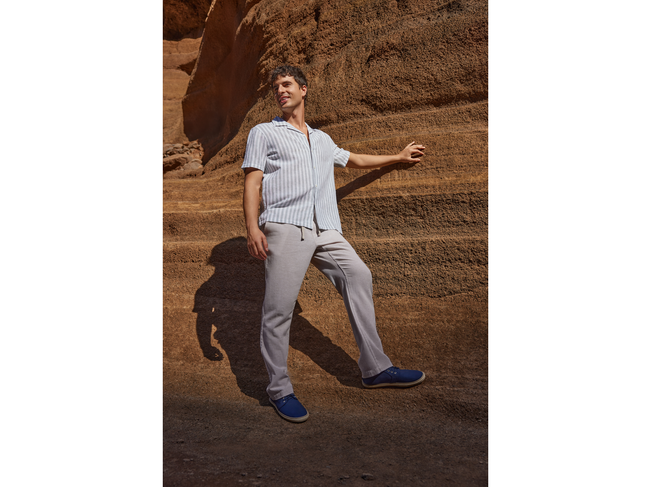 Man in striped short-sleeve shirt, light trousers, and blue espadrilles against a rock wall.