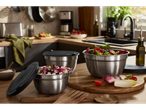 Bancada de cozinha com três tigelas de aço inoxidável com salada e tampas pretas, e utensílios de cozinha ao fundo.