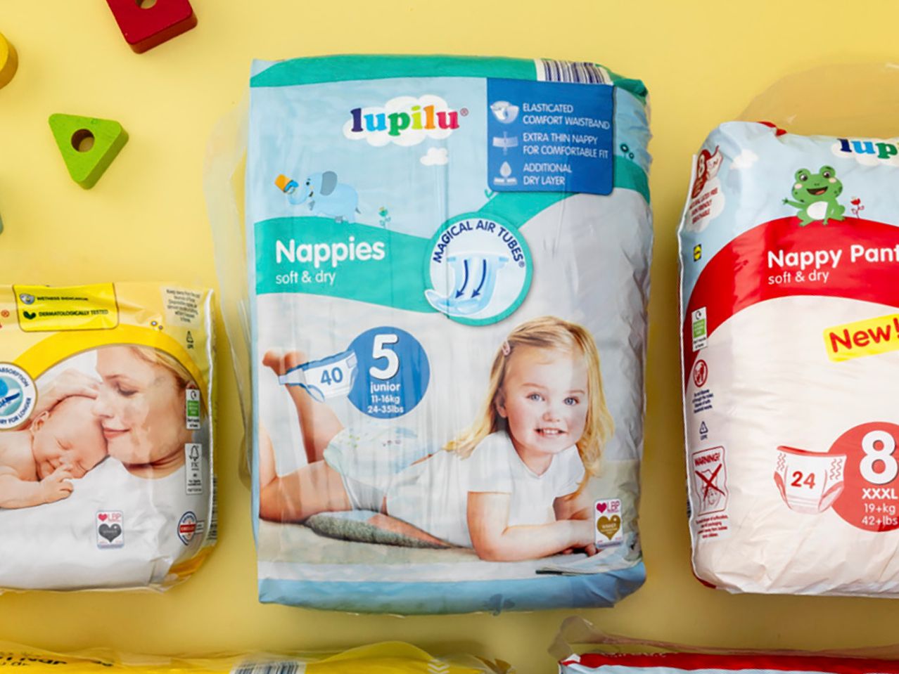 Three packages of Lupilu brand baby diapers and nappy pants on a yellow surface with wooden toys.