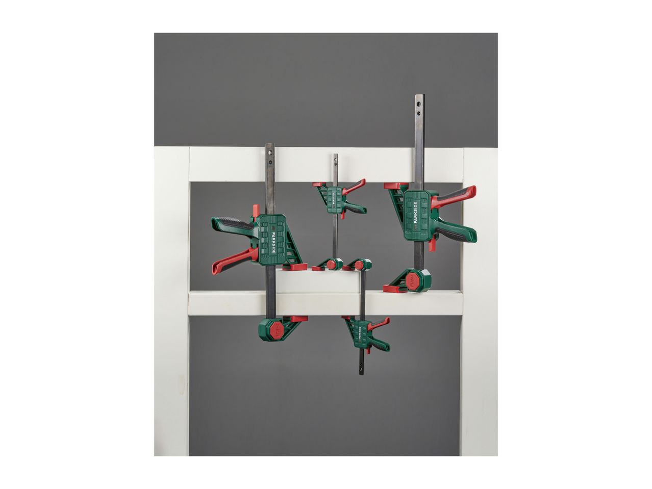A set of green and red bar clamps of various sizes, holding white wooden frames.