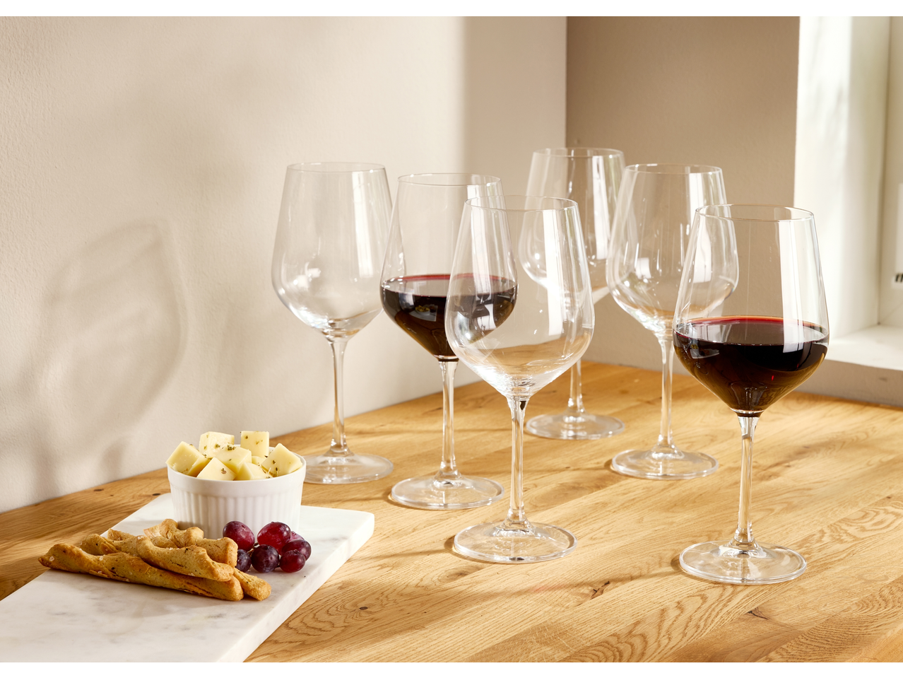 Red wine glasses, some filled, with cheese, grapes, and breadsticks on a wooden table.