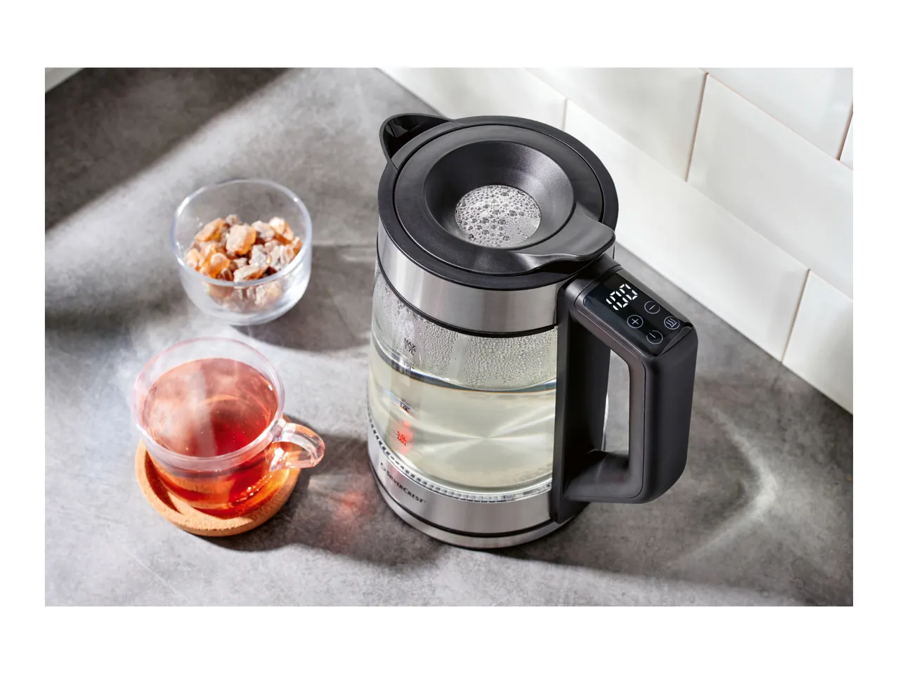 Silvercrest glass kettle with digital display, a cup of tea, and rock sugar on a counter.