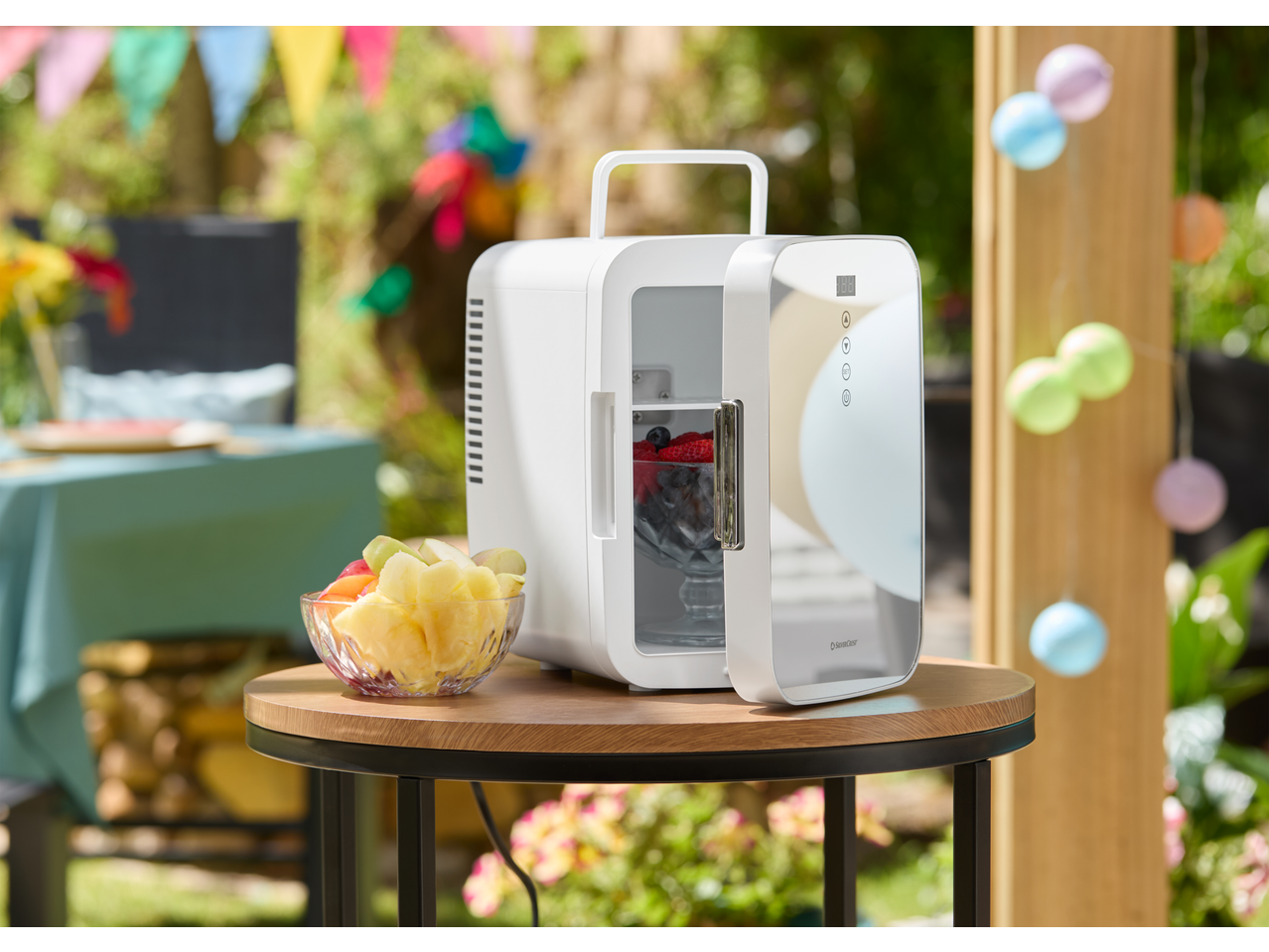 SILVERCREST® Mini Fridge with an open door revealing fruit, next to a bowl of fruit on a wooden table outdoors.