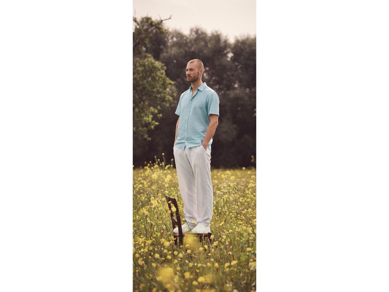 Uomo in camicia celeste e pantaloni bianchi in piedi su una sedia in un campo di fiori gialli.