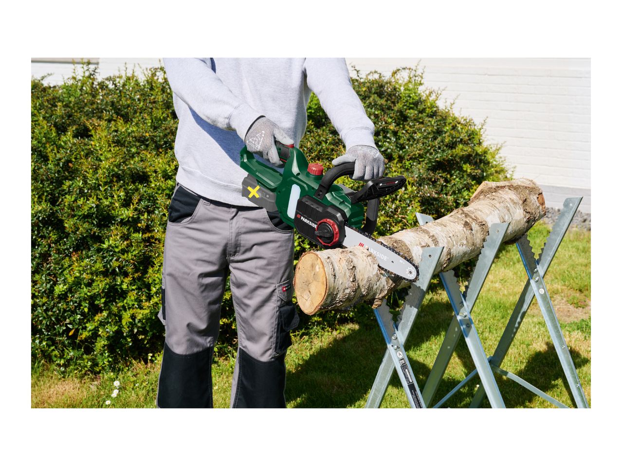 Person cutting a log with a cordless chainsaw on a sawhorse in a garden.