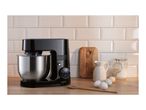 Black stand mixer with a stainless steel bowl on a wooden counter with baking ingredients.