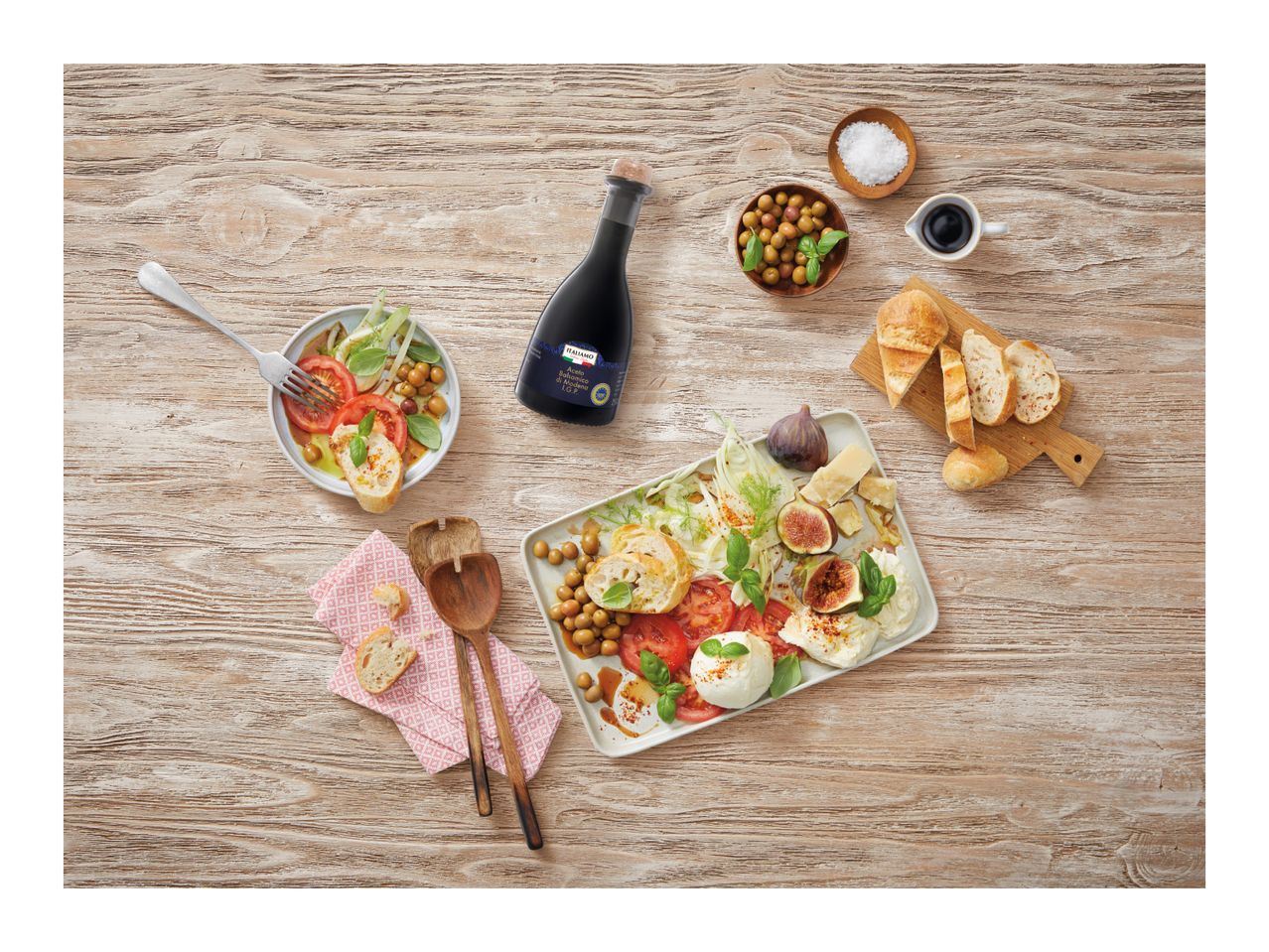 Balsamic vinegar bottle with a caprese salad, olives, figs, cheese, and bread on a wooden table.