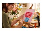 Woman painting flowers in a vase on a canvas, surrounded by art supplies and fresh flowers.