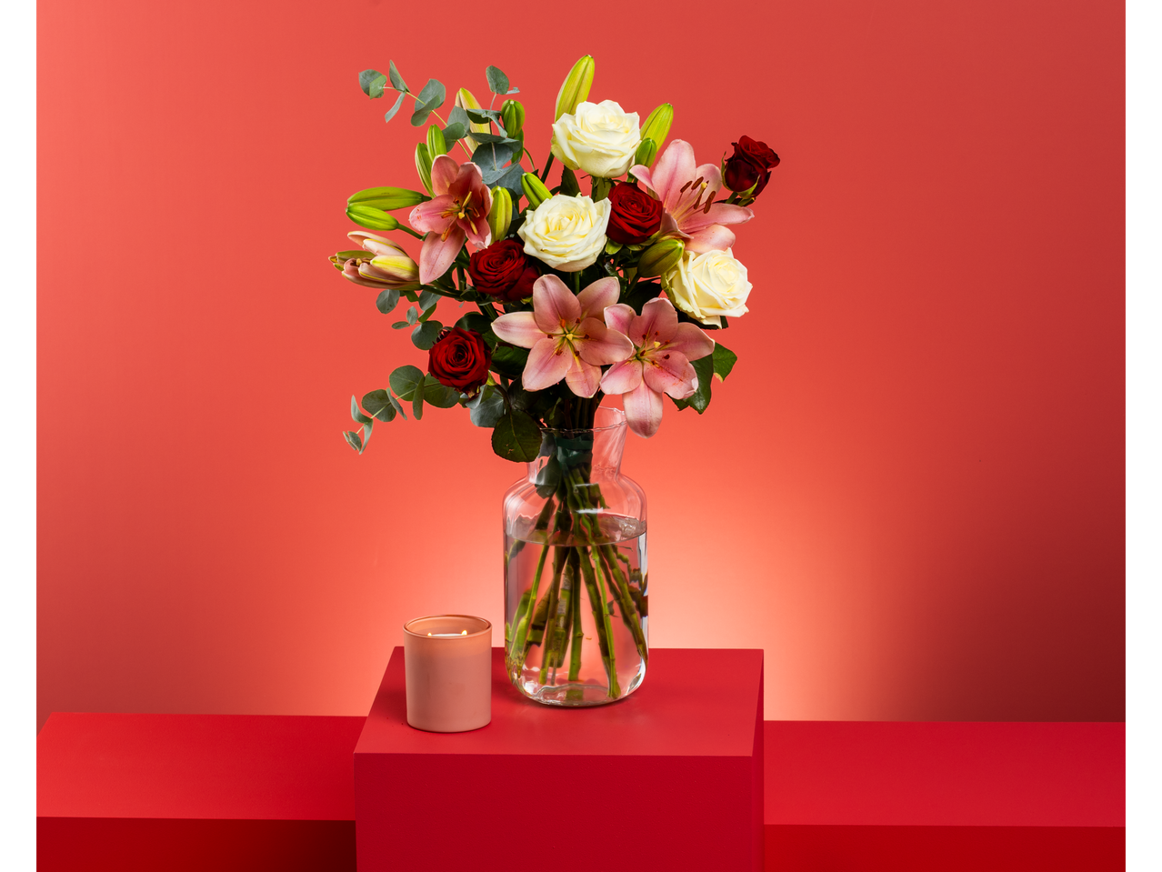 Bouquet of pink lilies, white and red roses, and eucalyptus in a vase with a candle.