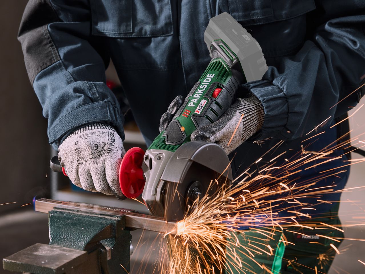 Person in workwear and gloves using an angle grinder to cut metal, creating sparks.