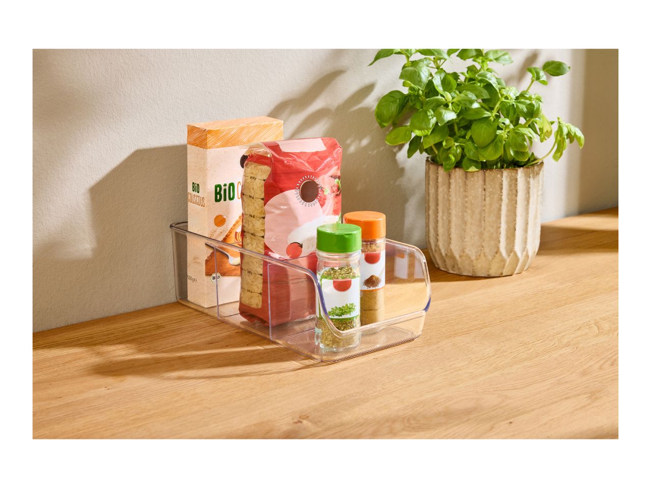 Clear organizer with couscous, rice, and spices on a wooden counter next to a basil plant.