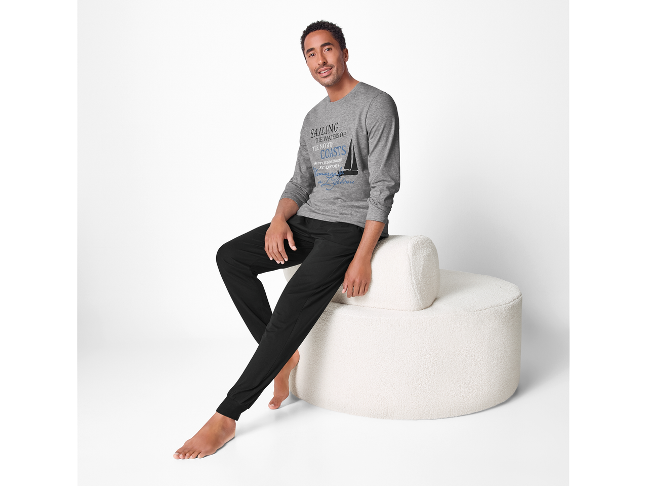 Moški v sivi majici z dolgimi rokavi z napisom „SAILING THE WATERS OF THE NORTH COASTS“ in črnih hlačah, sedi na belem kavču.