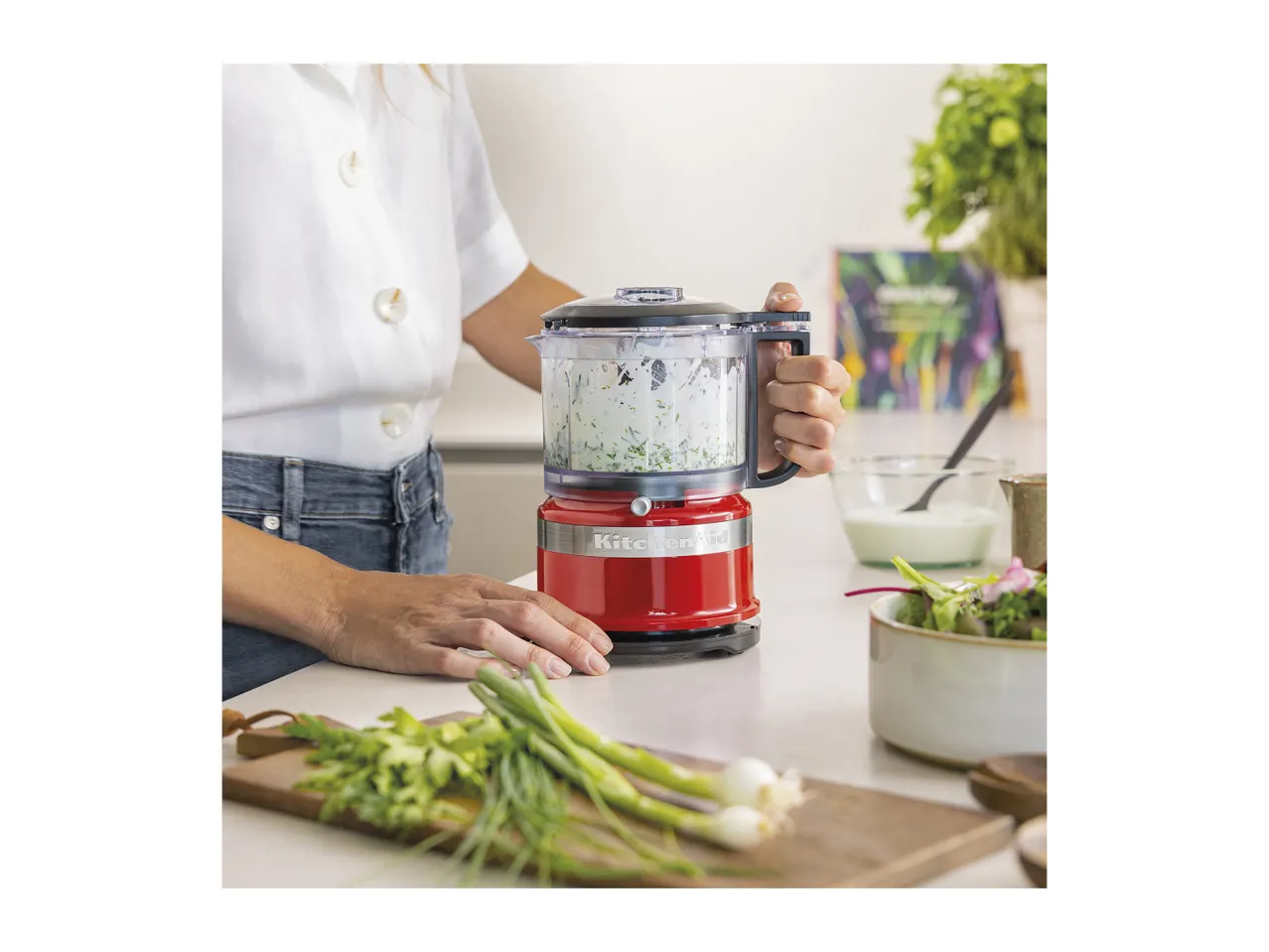 Person using a red food chopper to prepare a green herb sauce, with fresh vegetables nearby.