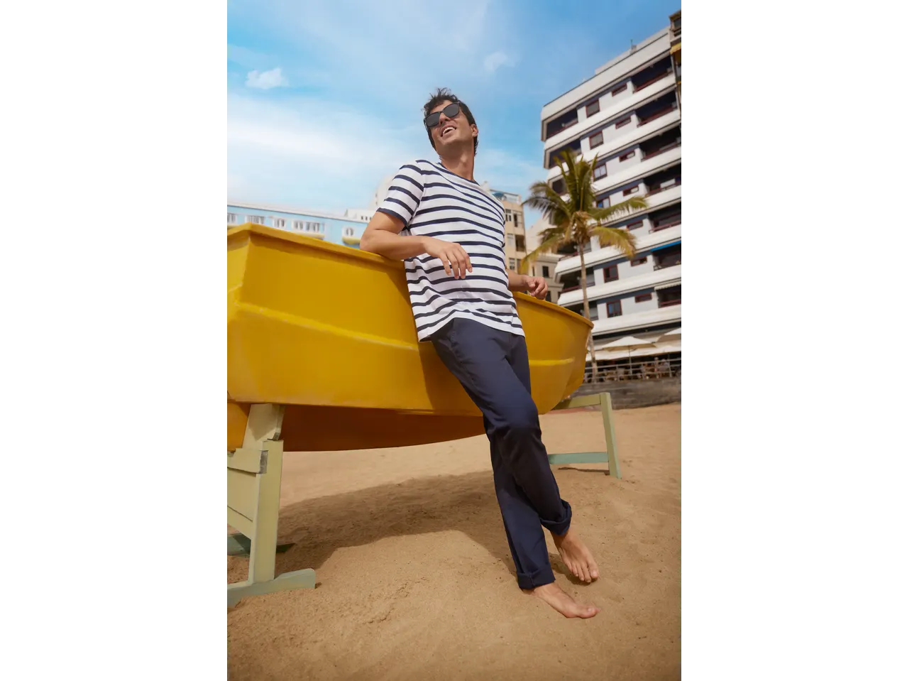 Muškarac u prugastoj majici ESMARA MEN i plavim pantalonama, naslonjen na žuti čamac na plaži.