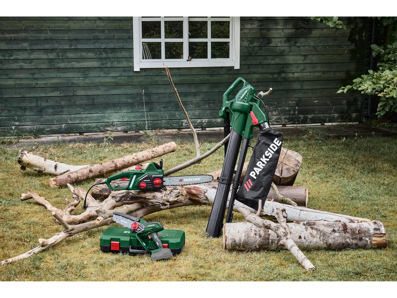 Uma motosserra elétrica, soprador de folhas e mini serra a bateria na grama, ao lado de toras.