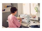 A woman using a white sewing machine at a desk near a window.