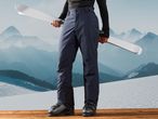 Person in dark blue ski pants and black top holding white skis, standing on a wooden deck.