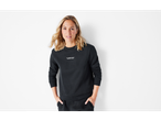 A woman in a black Esmara sweatshirt and sweatpants, hands in pockets, smiling.