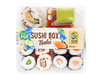 Škatla sušija z različnimi zvitki, omakami in napisom 'SUSHI BOX Toshi'.