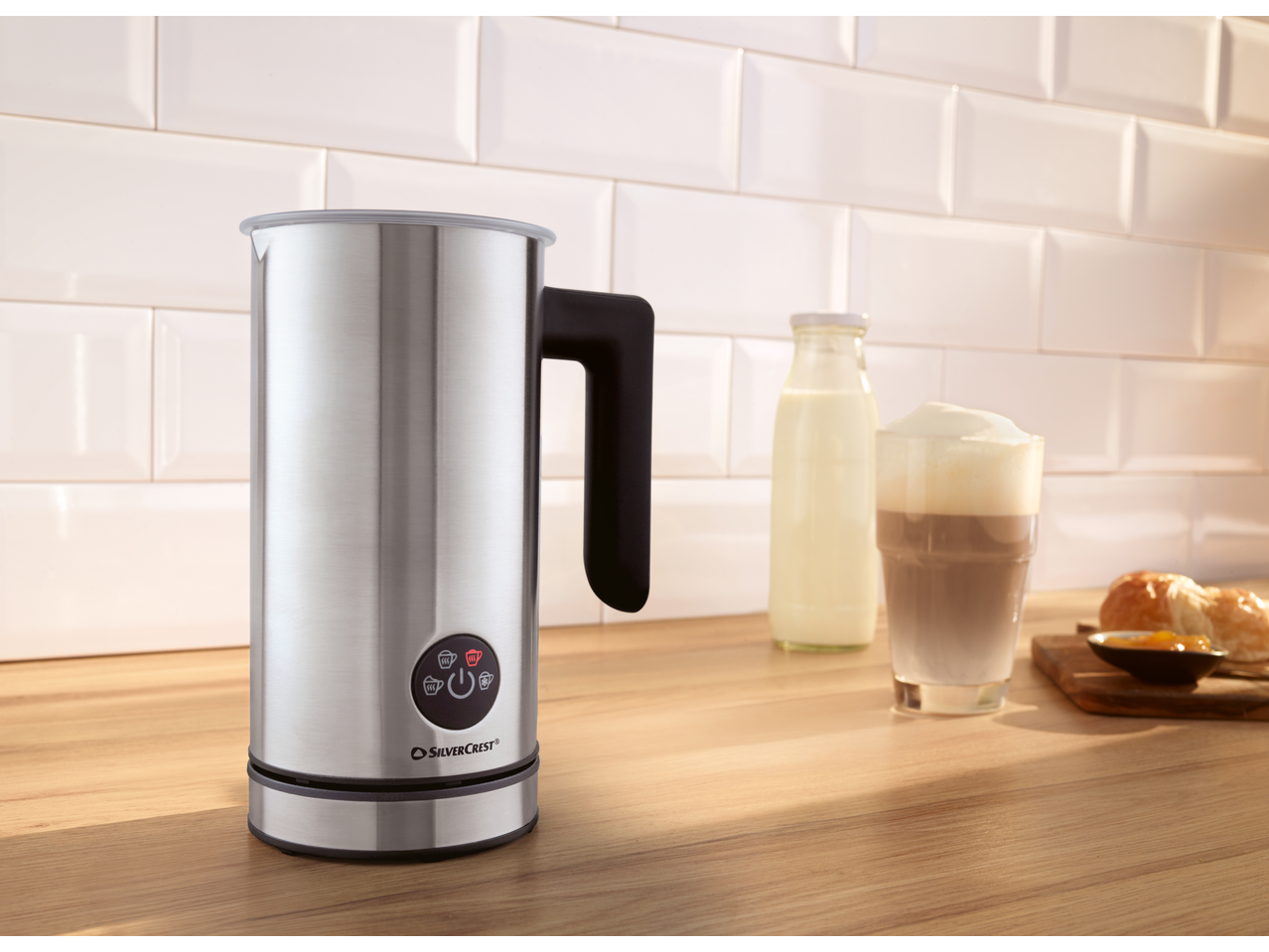 Stainless steel milk frother on a wooden counter with a latte and breakfast pastries.