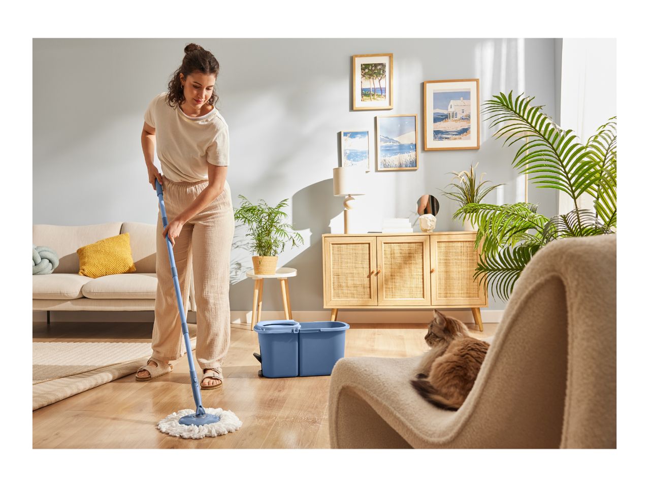 Woman mopping a wooden floor with a blue mop and bucket in a living room with a cat.
