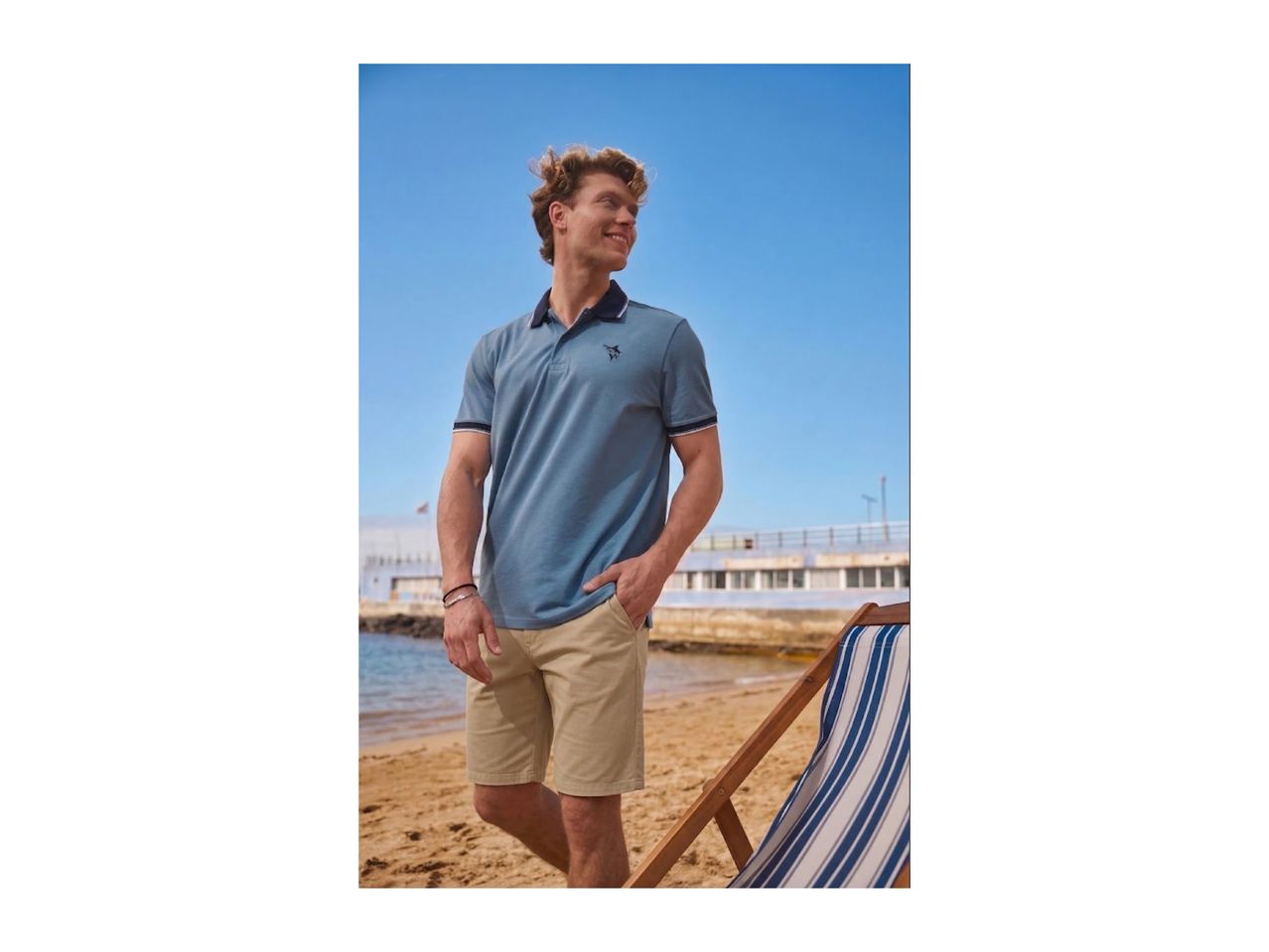 Man in blue polo shirt and beige shorts on a sandy beach with a striped deckchair.