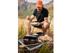 Man in CRIVIT sports top and orange cap eating outdoors with camping cooking gear.