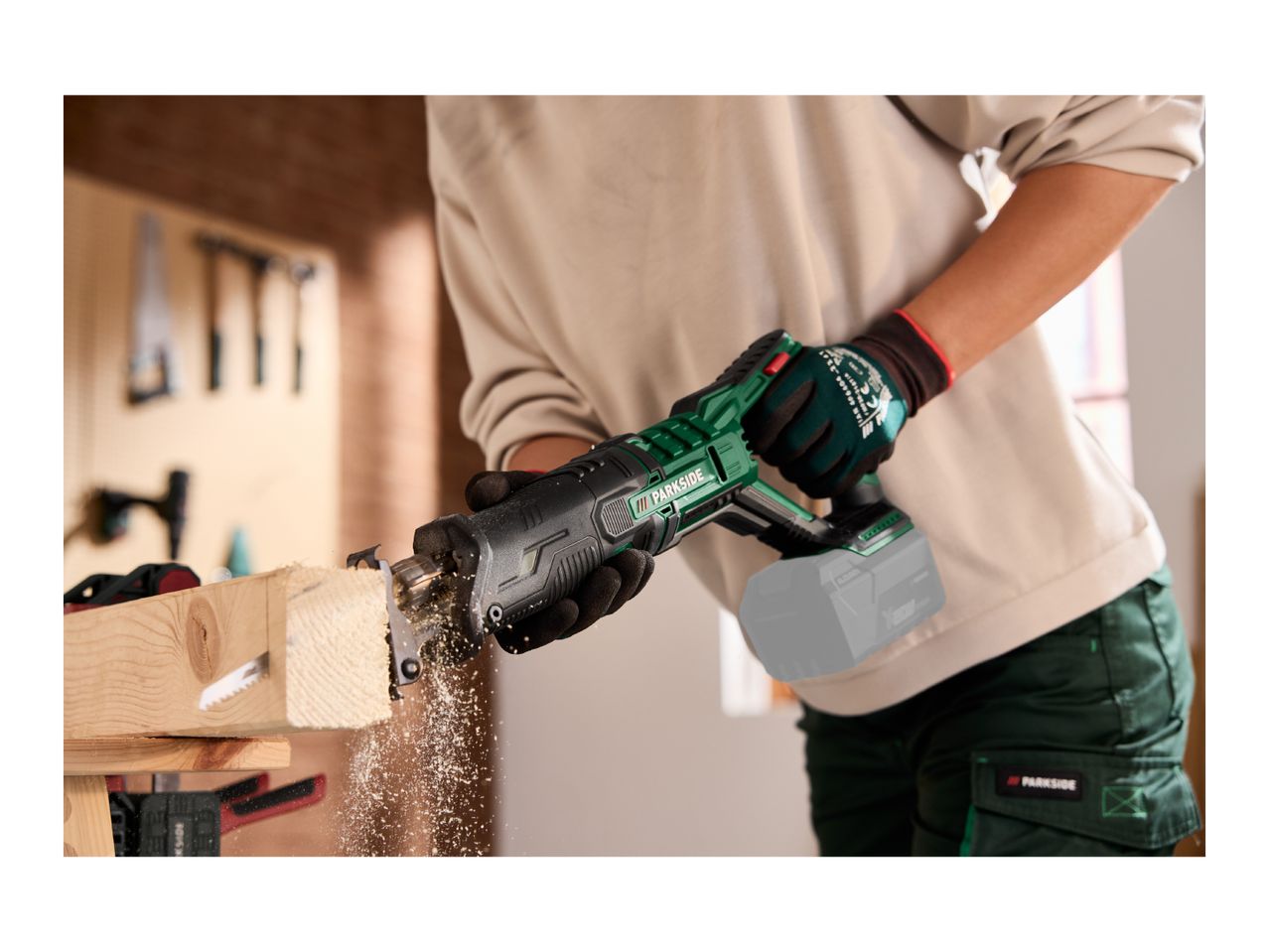 A person using a cordless reciprocating saw to cut a piece of wood in a workshop.