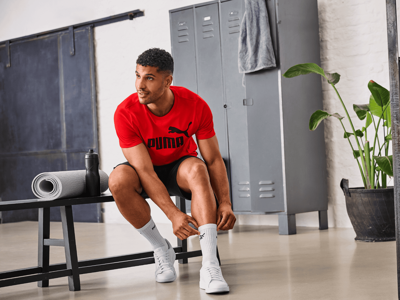 Mann in rotem T-Shirt und schwarzer Sporthose bindet sich die Schnürsenkel auf einer Bank.