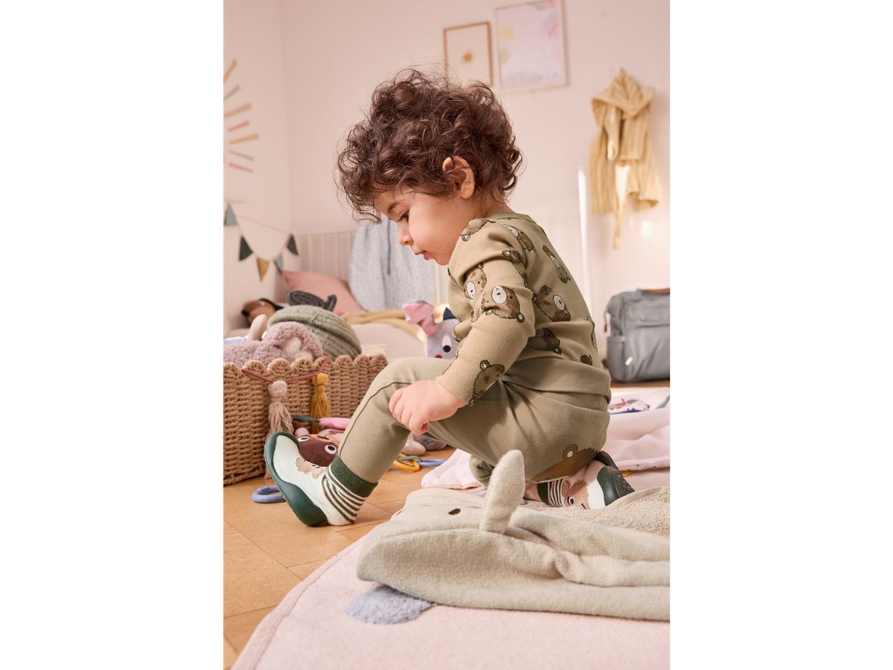 A toddler in a green bear-print outfit and animal-themed shoes plays on the floor.