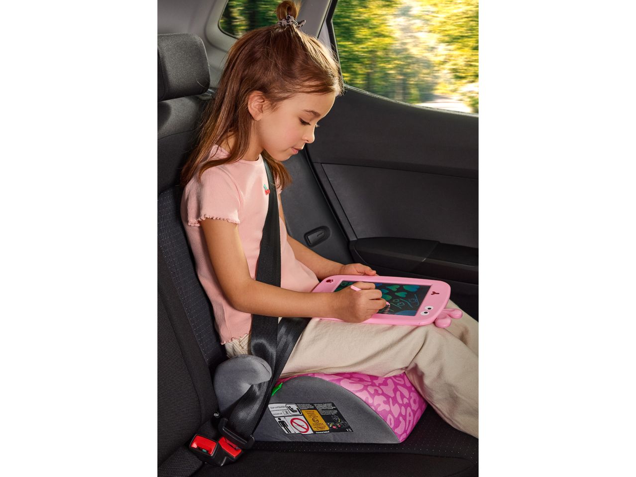 A child in a car booster seat drawing on a pink digital sketch pad.