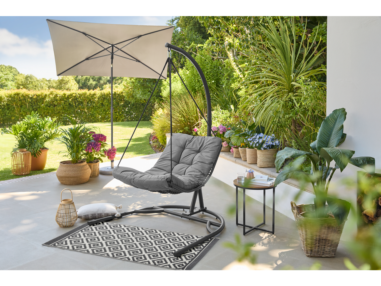 A garden swing chair with a grey cushion on a patio, surrounded by potted plants and an umbrella.