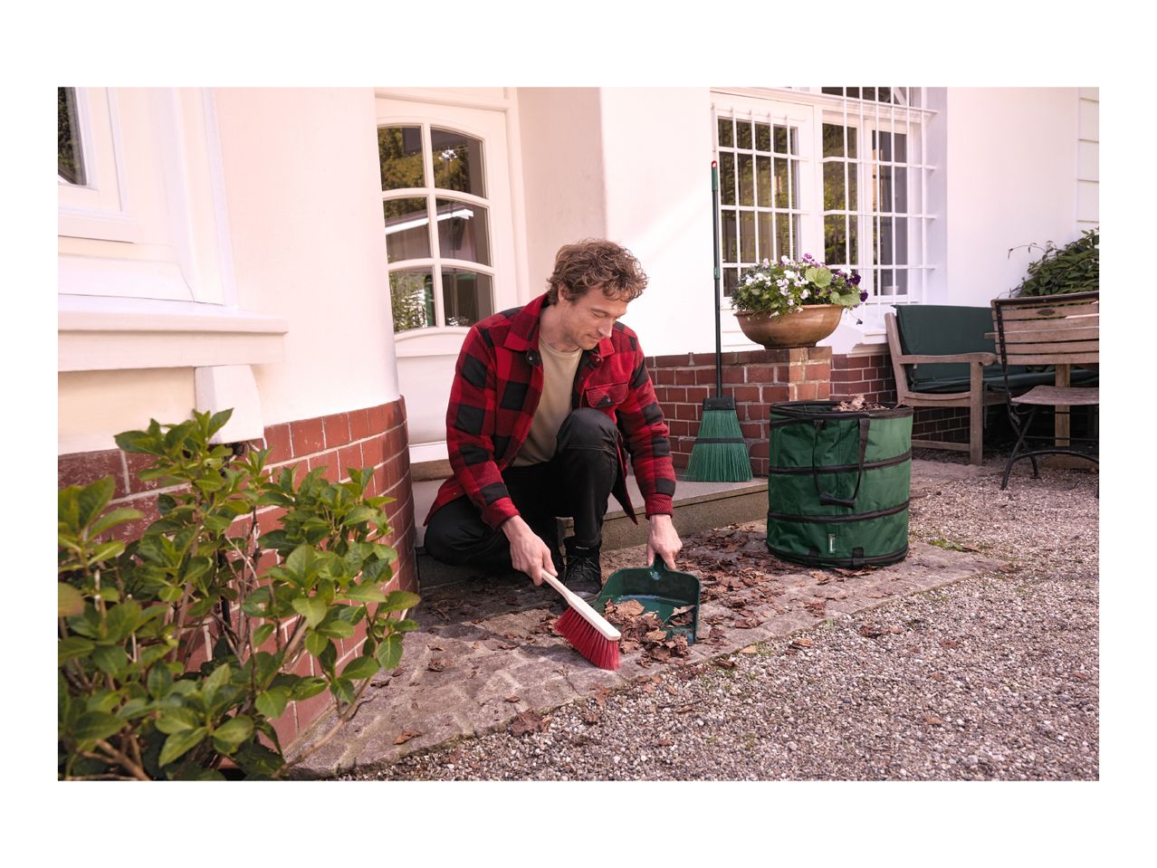 Man sweeping leaves into a green garden bag with a broom and dustpan.