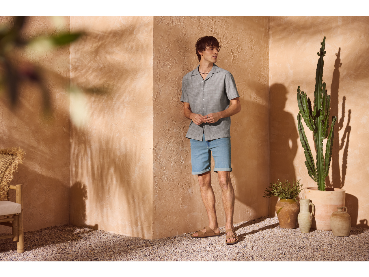 Homme en chemise grise à manches courtes et short en jean bleu clair, debout dans un coin ensoleillé avec un cactus.