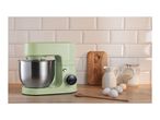 Green stand mixer with a stainless steel bowl on a wooden counter with baking ingredients.