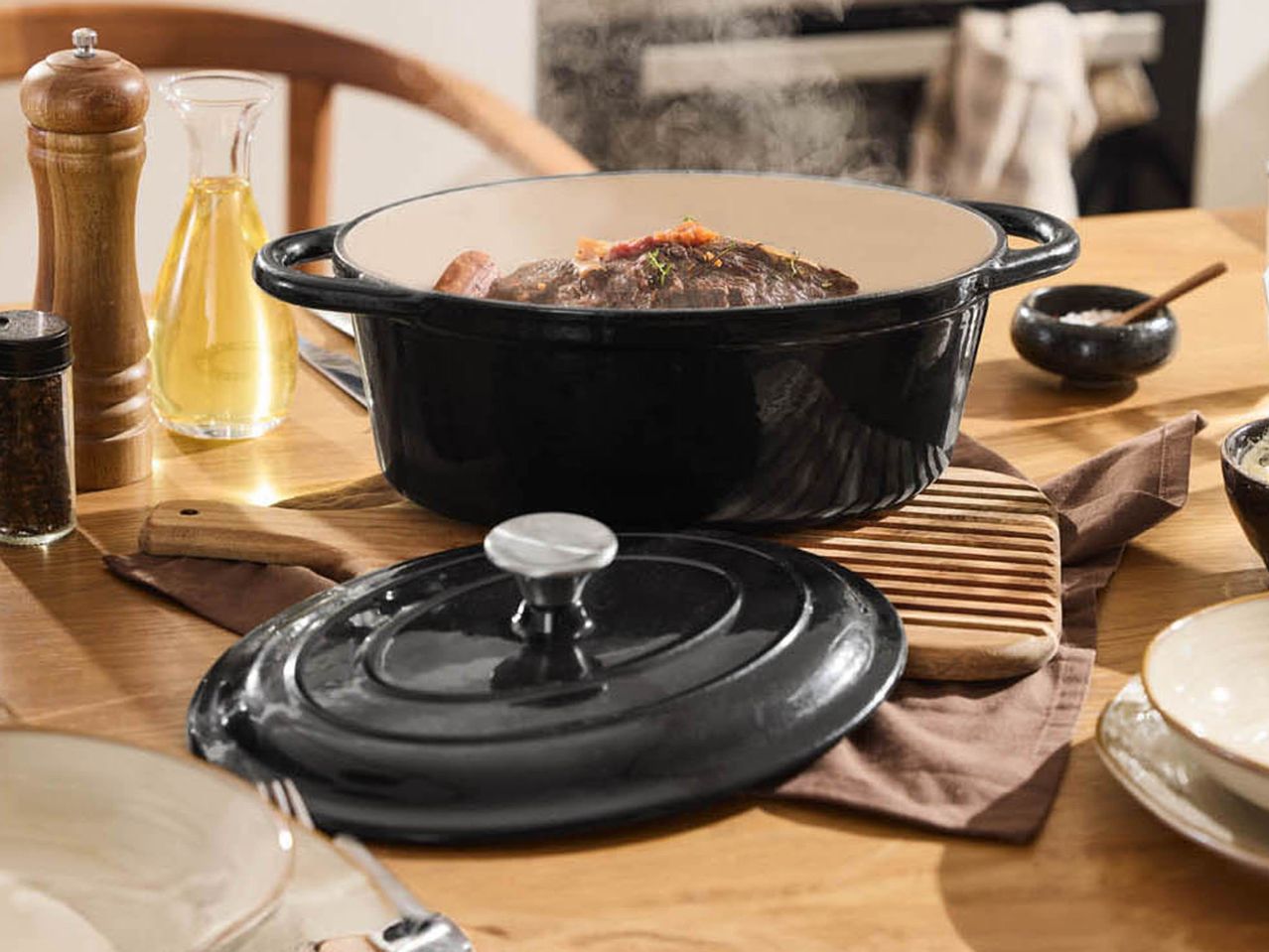 A black cast iron Casserole Dish with meat and sausage, surrounded by kitchenware on a wooden table.