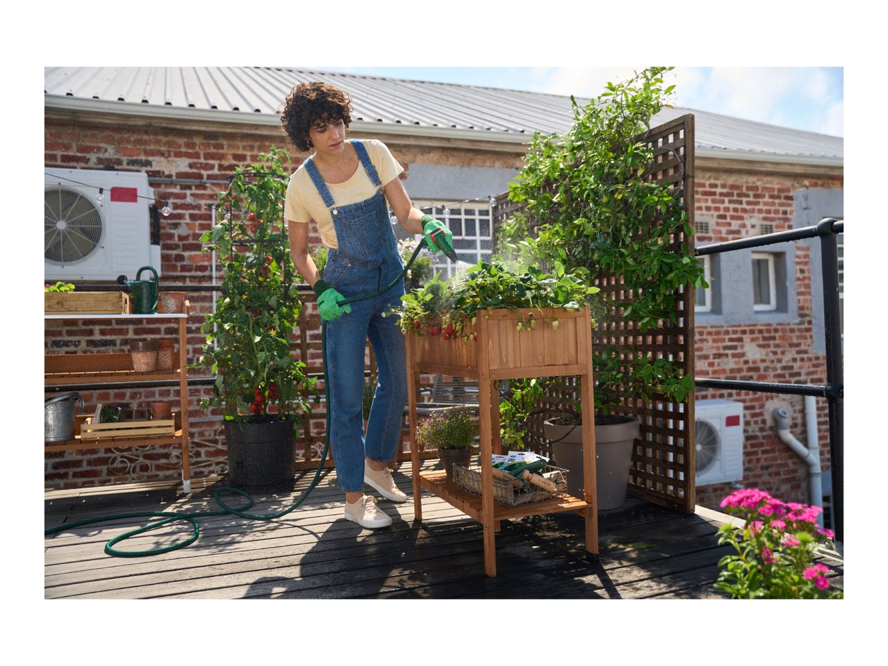 Woman watering plants in a raised wooden planter on a balcony garden.