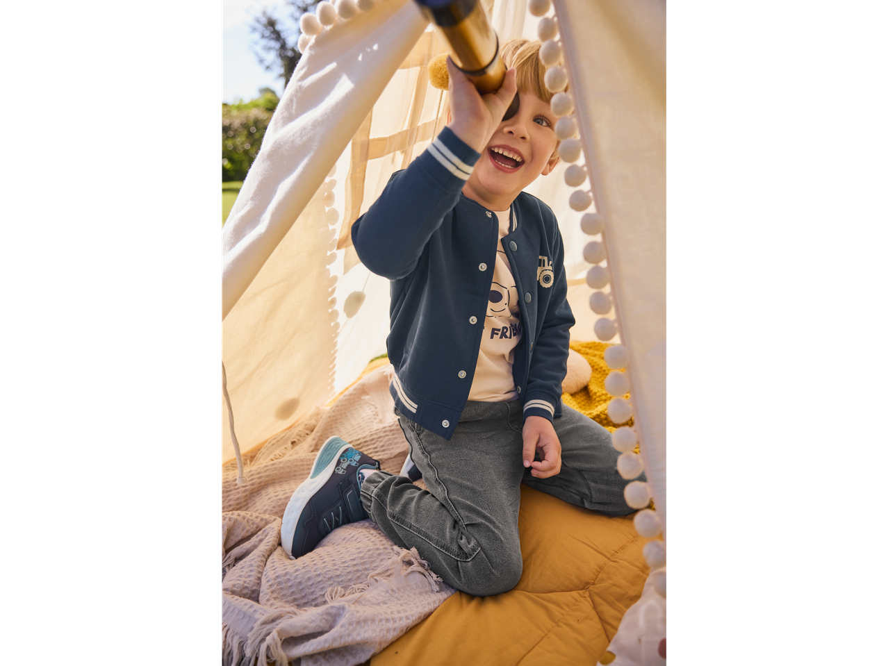 A boy plays with a telescope inside a teepee wearing a navy blue bomber jacket and grey jeans.