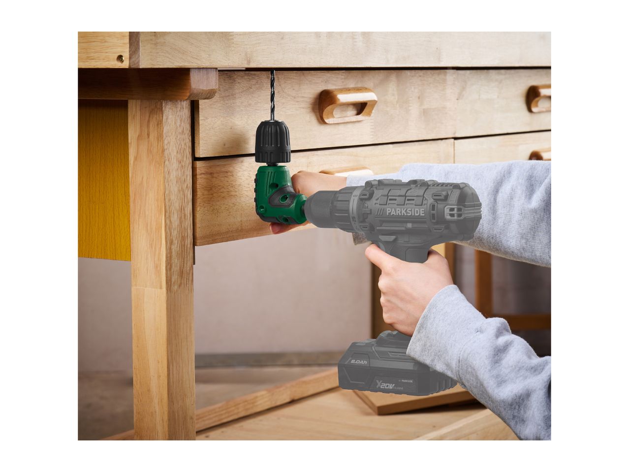 A cordless drill with a right-angle attachment drilling into a wooden workbench.
